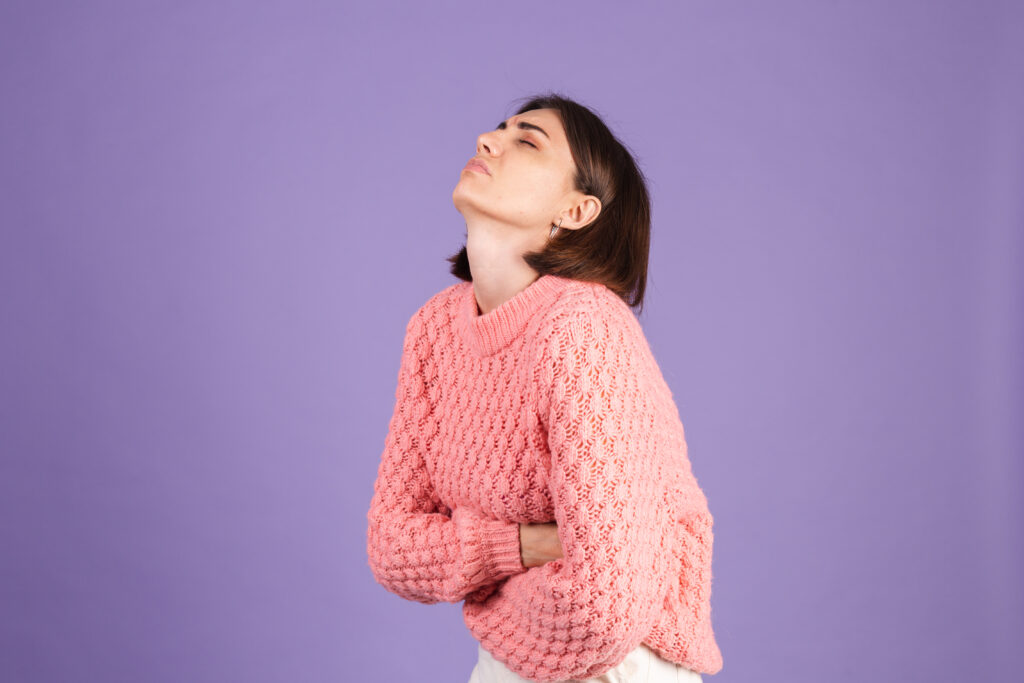 young brunette in pink sweater isolated on purple background suffering stomach ache with painful grimace, feeling sudden period cramps, gynecology concept